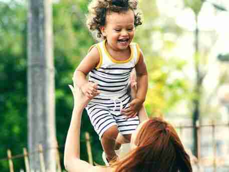ADHD Essentials: 8 Therapist-Guided Tips for Thriving with Your ADHD Brain 11 A person lifts a smiling child in a striped outfit high into the air outdoors, capturing a moment of joy and playfulness. Amidst the blurred greenery and visible fence, these carefree moments serve as essential breaks in a world that often demands attention.